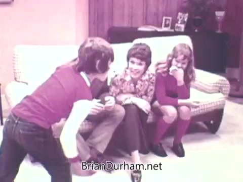 The still from 'Ideal 'Up Set' game - 1973' features three children interacting in a living room setting. One boy is animatedly gesturing towards a seated girl and woman, suggesting a playful or engaging moment. The room has a cozy, vintage aesthetic typical of the 1970s, with a patterned couch and wooden paneling visible in the background.