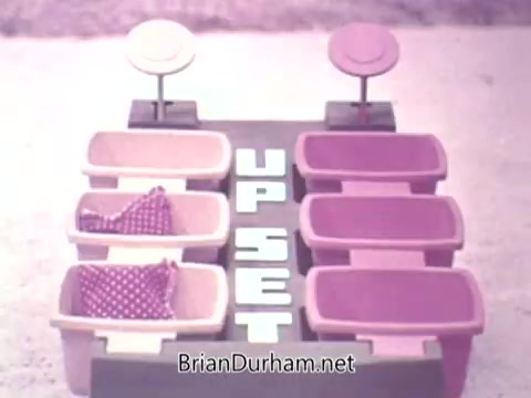 The still depicts a game setup from the 1973 Ideal 'Up Set' game. It features several colored containers arranged in two rows, with pink and light purple hues. In the center, the words 'UP SET' are prominently displayed. Some containers contain polka-dotted fabric, suggesting a playful, possibly competitive atmosphere. The overall aesthetic looks retro, typical of 1970s game design.