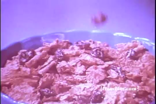 The still from the 1970s Kellogg's Raisin Bran commercial features a close-up of a bowl filled with crunchy flakes and plump raisins. The image captures a moment where a raisin is seen falling into the bowl, emphasizing the cereal's appeal and the inclusion of raisins. The overall color palette has a nostalgic tone typical of vintage commercials, with an inviting and appetizing presentation.