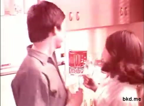 The still from the 'Duz Soap TV Commercial' features two individuals in a kitchen setting. The scene is likely lighthearted, with one person gesturing playfully towards the other. A prominently displayed Duz soap box is visible in the background, suggesting the product's importance in the commercial. The lighting has a vintage tone, typical of 16mm film, creating a nostalgic feel.