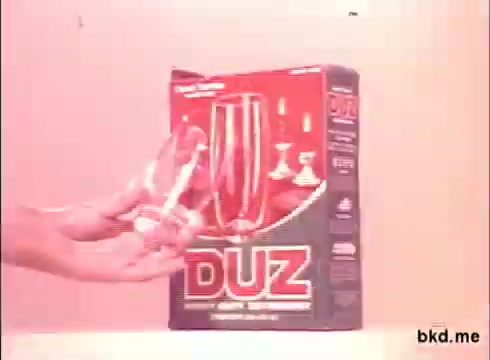 The still features a hand holding a glass next to a prominently displayed box of Duz soap. The packaging is red with bold white lettering, emphasizing the brand name. The glass appears to be clean and potentially highlights the product's effectiveness in dishwashing or cleaning. The background is softly lit, enhancing the focus on the product.