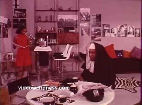 The still from *Come Back, Charleston Blue* features a scene set in a stylish, modern apartment reflecting the blaxploitation aesthetic of the early 1970s. In the foreground, a woman in a red dress stands, possibly conversing or interacting with a seated character. The seated character, dressed in a nun's habit, appears engaged, adding a layer of intrigue to the scene. The decor includes modern furnishings and various art pieces on the walls, capturing the cultural vibe of the time. The film explores themes of justice and community, following the characters Coffin Ed Johnson and Grave Digger Jones as they navigate crime and morality in Harlem.