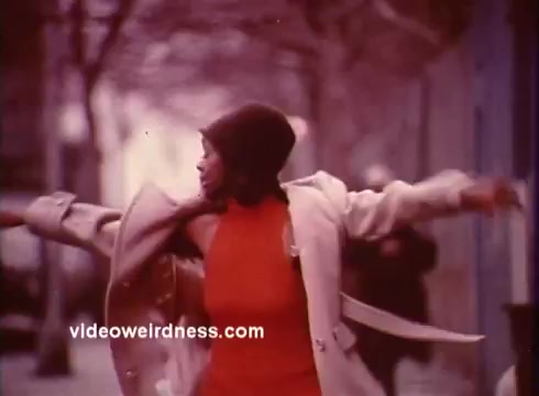 The still from 'Come Back, Charleston Blue' captures a vibrant moment with a woman in a striking red dress and cream coat, exuding confidence and freedom as she twirls in an urban setting. The background is blurred, suggesting motion, emphasizing her dynamic presence against a cityscape. This scene reflects the film's themes of resilience and cultural identity within the blaxploitation genre, connecting to the broader narrative of African American experiences in the early 1970s. The film, featuring characters Coffin Ed Johnson and Gravedigger Jones, weaves together elements of crime, humor, and social commentary.