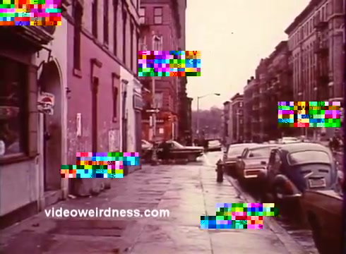 The 16mm still from 'Come Back, Charleston Blue' captures a city street scene, likely set in a 1970s urban environment. The image shows a wet pavement, suggesting recent rain, with vintage cars lined along the curb. Buildings feature brick facades, hinting at a bustling neighborhood atmosphere. This scene encapsulates the blaxploitation genre's aesthetic, focusing on urban life and cultural narratives, reflecting the struggles and stories of characters like Coffin Ed Johnson and Gravedigger Jones, who navigate the complexities of their environment.