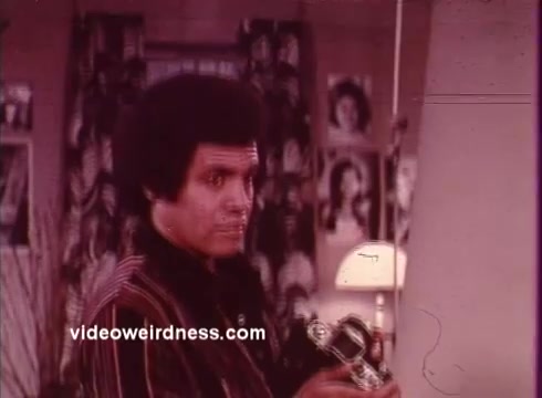 The still from 'Come Back, Charleston Blue' features a character with a distinct afro hairstyle, holding a vintage camera. The setting appears to be a room with patterned wallpaper and a lamp, adding to a retro ambiance. This film, part of the blaxploitation genre, showcases themes of urban life and crime, centered around the characters Coffin Ed Johnson and Gravedigger Jones, portrayed by Godfrey Cambridge and Raymond St. Jacques. The film addresses socio-political issues while providing entertainment through its unique style and storytelling.