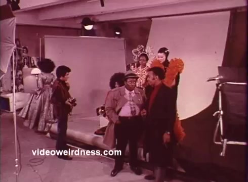 The still from 'Come Back, Charleston Blue' features a bustling scene indicative of the film's vibrant blaxploitation style. In the foreground, two male characters engage in conversation, dressed in stylish attire reflective of the era. Behind them, several women showcase elaborate costumes, adding to the film's artistic flair. The backdrop suggests a photography or film set, with lighting equipment that emphasizes the cinematic context. The overall composition captures the film's themes of community and cultural expression prevalent in the 1970s, alongside the humor and social commentary characteristic of Chester Himes' work.