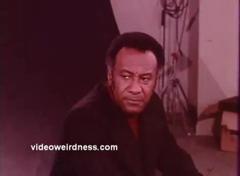The still from 'Come Back, Charleston Blue' features a serious-looking man in a black outfit, conveying a sense of intensity and intrigue. He appears to be in a studio setting, with studio equipment visible in the background. The film explores themes of crime and justice, centered around the characters Coffin Ed Johnson and Gravedigger Jones, who navigate the complexities of their environment in a gritty urban backdrop. The visual style reflects the blaxploitation genre's emphasis on character depth and social commentary.