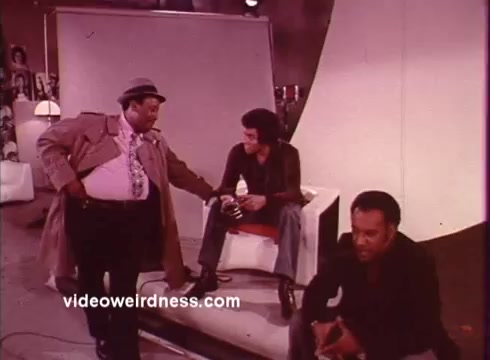 The still from *Come Back, Charleston Blue* features three characters in a stylized, presumably film set environment. One character, dressed in a trench coat and fedora, stands with an assertive posture, while another sits on a white chair, holding a cup. The third character is seated on the ground, looking contemplative. The scene captures the blaxploitation aesthetic, emphasizing urban themes and the camaraderie among characters. The film, connected to Chester Himes' work, explores themes of crime and community, typical of the genre's narrative style.