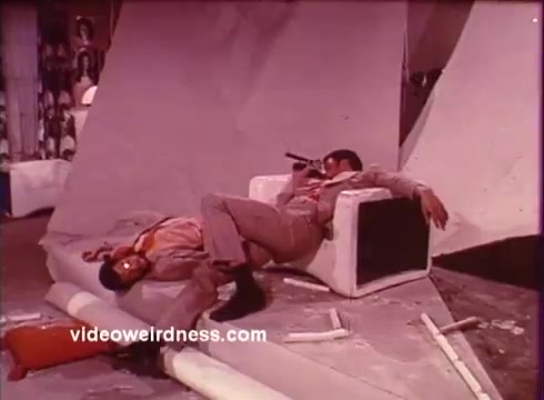 The still from 'Come Back, Charleston Blue' features two characters in a dramatic pose, capturing the essence of the blaxploitation genre. One character reclines lazily on a white chair, holding a microphone, while the other appears to be slumped nearby, suggesting a tense or relaxed moment within a stylized setting. The color palette and composition reflect the film's 1970s aesthetic, emphasizing a blend of humor and social commentary typical of Chester Himes' work. This scene likely highlights themes of camaraderie and the gritty realities faced by the protagonists, Coffin Ed Johnson and Gravedigger Jones, as they navigate their world.