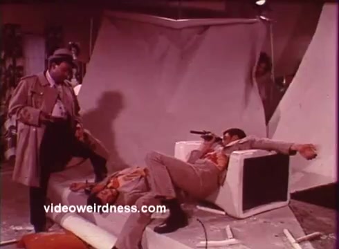 The still from 'Come Back, Charleston Blue' portrays a tense scene typical of the blaxploitation genre. In the foreground, one character is dramatically lying on a sofa, appearing injured or incapacitated, while another stands nearby, possibly in a stylish trench coat, suggesting a sense of urgency or confrontation. The setting is chaotic, with disordered elements like a torn backdrop, enhancing the intense atmosphere of the scene. This imagery reflects the film's themes of crime, survival, and the complex dynamics within the African American community during this era, as inspired by Chester Himes' work.