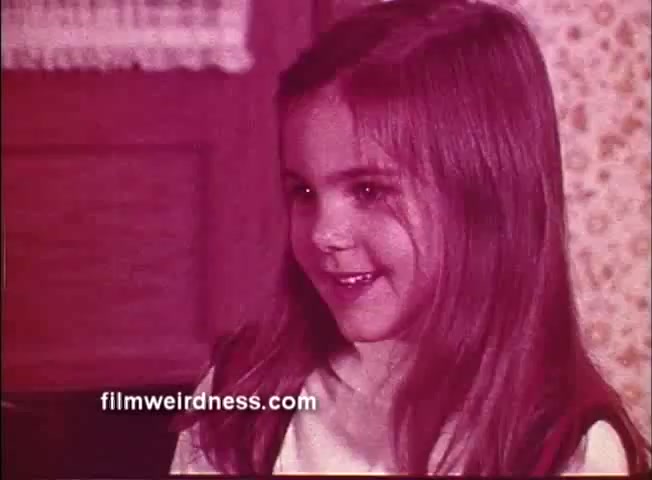 The still features a young girl with long hair, smiling and appearing engaged in conversation. The color palette has a warm, reddish tint, contributing to a nostalgic atmosphere. The setting suggests a cozy indoor environment, typical of children's programming aimed at younger audiences. This scene likely showcases playful interaction, reflecting the lighthearted tone of 'How's The Weather, Lucy - Colorforms Peanuts.'