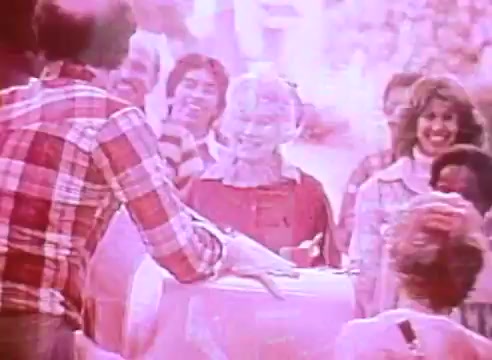 The still from 'Welcome to Fulton St' - energy efficient houses PSA 2 features a group of diverse individuals, including a woman in a red outfit and several others in casual attire, gathered around a man in a checkered shirt. The scene radiates a sense of community and engagement, likely celebrating energy-efficient housing. Bright, soft lighting gives the image a warm, inviting feel.