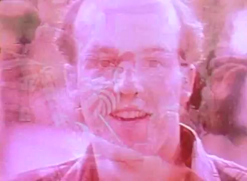 The still features a close-up of a smiling man, with a soft, pink hue overlay creating a dreamlike effect. The blurred background suggests a lively atmosphere, possibly hinting at a community-focused theme around energy-efficient housing. The imagery conveys optimism and warmth, aligning with the message of sustainable living in 'Welcome to Fulton St.'