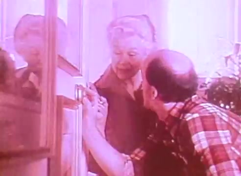 The still from 'Welcome to Fulton St - energy efficient houses PSA 2' features a man and an older woman engaged in a conversation at a door. The man, wearing a plaid shirt, seems to be explaining something, while the woman looks attentive and approachable. The warm lighting and soft colors suggest a cozy atmosphere, typical of community-focused initiatives promoting energy efficiency in homes.