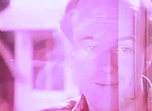 The still features a smiling man in a plaid shirt, framed partially by a door. There’s a soft, pinkish hue affecting the image, creating a warm, inviting atmosphere. The background suggests a home setting, likely connected to themes of energy efficiency and comfort in housing, fitting the context of a public service announcement.