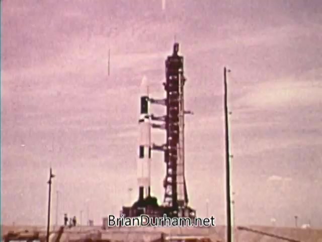 The still features a rocket on a launch pad with a support tower beside it, set against a cloudy sky. The image likely conveys themes of exploration and technology, relevant to the 1976 Bicentennial PSA. The color tone suggests a vintage look, characteristic of 16mm film.