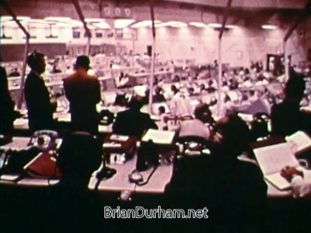 The still from 'Horizons On Display - Bicentennial PSA from 1976' features a busy control room setup, likely related to space or aerospace operations. A group of men in suits and headset telephones are engaged in communication, while others analyze documents and data. The environment suggests a high-stakes, collaborative effort typical of mission control centers, reflecting technological progress during the bicentennial period. The overall aesthetic conveys a sense of urgency and seriousness associated with scientific endeavors.