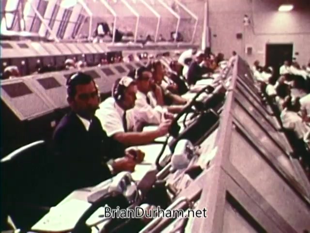 The still from 'Horizons On Display - Bicentennial PSA' features a busy control room with multiple operators, dressed in white shirts and headsets, intently monitoring and managing various instruments. The atmosphere reflects a sense of urgency and focus, indicative of a significant event or operation, likely related to space or technology during the 1976 Bicentennial period. The room is equipped with numerous dials and screens, emphasizing the high level of coordination required for the task at hand.