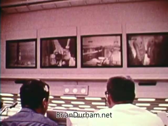 The still captures two men seated in a control room, focused on multiple screens displaying live footage related to space missions. The environment suggests a high-tech setting, indicative of the 1970s space exploration era. Each screen shows different scenes, possibly of rocket launches or mission operations, emphasizing the technological advancements and national pride during the Bicentennial celebrations in 1976.