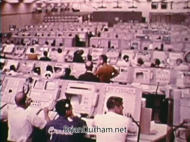 The still from 'Horizons On Display - Bicentennial PSA' features a large control room filled with numerous operators at their stations. Each station is equipped with various control panels and monitors. The environment is busy, showcasing a team of workers focused on tasks, likely related to aerospace or scientific operations, reflecting the technological advancements of the time. The overall atmosphere suggests a sense of collaboration and dedication to progress.