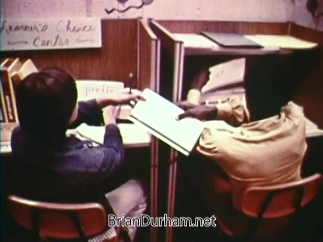 The still from 'Horizons On Display - Bicentennial PSA from 1976' features two students engrossed in their work at a study area. One student, dressed in a blue jacket, is pointing to a page in a book, while the other, wearing a light-colored shirt, is looking at the open book. In the background, a sign indicates a 'Career Choice Center,' and there are bookshelves with additional materials. The scene conveys a sense of collaboration and educational engagement.