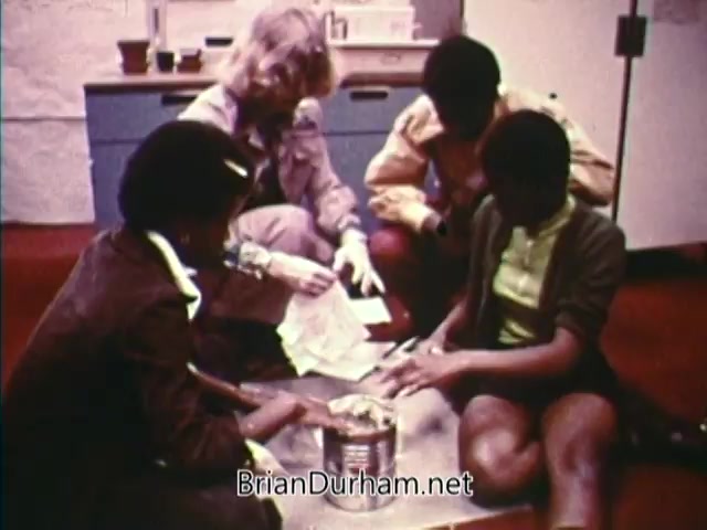 The still from 'Horizons On Display - Bicentennial PSA' features a group of four individuals, including two young women and one adult, engaged in an activity on the floor. They are seated around a table with a tin can and papers. The environment appears to be educational, likely a classroom or workshop setting. Emphasis is on collaboration and learning.