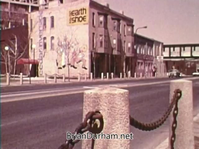 The still from 'Horizons On Display - Bicentennial PSA' features a street scene with a chain-link fence in the foreground and a weathered building displaying a mural for 'Earth Shoe.' The atmosphere has a vintage feel, indicative of the mid-1970s. Notable elements include the urban architecture and the emphasis on community aesthetics during the Bicentennial era.