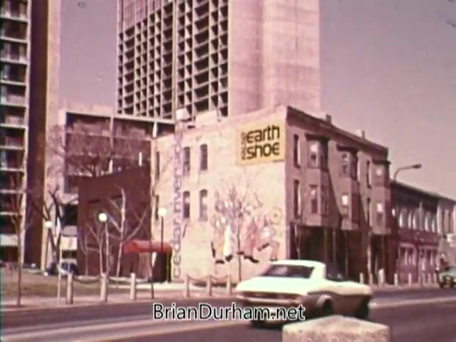 The still from 'Horizons On Display - Bicentennial PSA' features a street scene showcasing a mix of urban architecture. A partially demolished older building displays a mural and an advertisement for 'Earth Shoe.' In the foreground, a white car drives along the road, while modern high-rise buildings loom in the background. The overall aesthetic reflects a mid-1970s urban landscape.