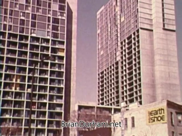 The still from 'Horizons On Display - Bicentennial PSA' features a view of urban architecture, highlighting two large high-rise buildings. The left structure displays a grid of balconies, while the right building appears more streamlined with a modern design. In the foreground, a sign for 'earth shoe' is visible on a lower building, suggesting a commercial presence. The scene conveys a sense of urban development typical of the 1970s, with a muted color palette.