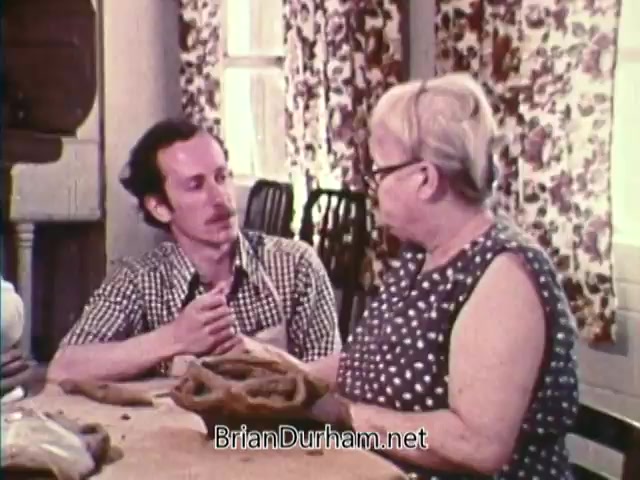 The still depicts a seated man and woman engaged in conversation at a table. The man, dressed in a checked shirt, appears to be attentively listening or explaining something, while the woman, wearing a polka-dotted dress and glasses, is holding an object, possibly a piece of pottery or a craft item. The room features floral curtains and a warm, homey atmosphere, suggesting a focus on community and heritage. This scene is part of a Bicentennial public service announcement from 1976, highlighting themes of cultural preservation and storytelling.