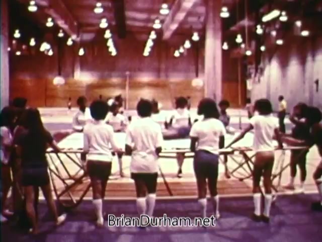The still from 'Horizons On Display - Bicentennial PSA' features a group of young individuals, mostly dressed in white shirts and athletic shorts, working together around a table or display in a gymnasium setting. The atmosphere suggests collaboration and community involvement, emblematic of the focused efforts during the 1976 Bicentennial celebrations. The background showcases a well-lit sports facility, emphasizing a sense of activity and engagement.
