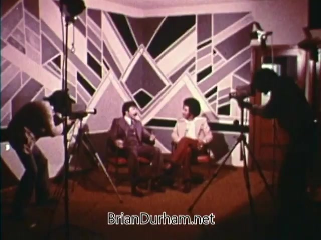 The still features two men seated in a stylized interview setting, surrounded by geometric wall art. They appear to be engaged in a conversation, with cameras pointed at them from both sides. The scene conveys a vintage aesthetic typical of 1970s public service announcements. The overall atmosphere suggests a moment of dialogue or reflection, aligning with the themes of awareness and community featured in 'Horizons On Display.'