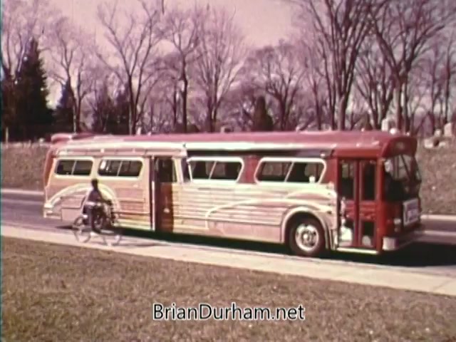 The still features a vintage red and silver bus parked along a road, with a cyclist approaching it. The backdrop includes bare trees, suggesting early spring or late fall, and a serene atmosphere typical of the 1970s. The image evokes themes of transportation and community, fitting for a public service announcement from the Bicentennial celebrations in 1976.