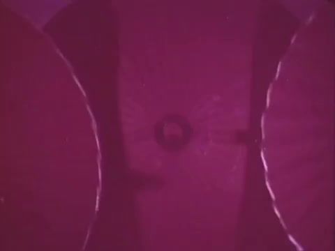 The still features a vibrant, pink background with circular elements that create a sense of movement and style. The design is likely intended to capture attention and evoke a playful, energetic vibe typical of a hairspray commercial. The overall aesthetic suggests glamour and creativity, aligning with the promotion of hair products.