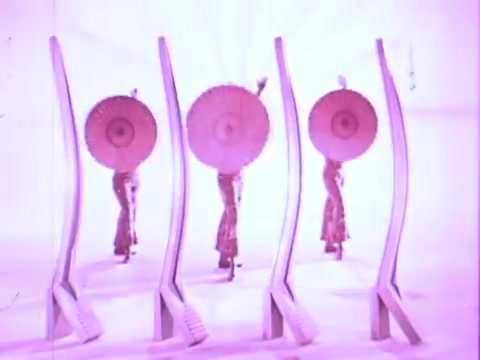 The still features four dancers in vibrant outfits, each holding large, round umbrellas. They are positioned against a soft pink background, flanked by stylized, elongated hair products. The overall aesthetic is playful and glamorous, characteristic of a retro commercial.