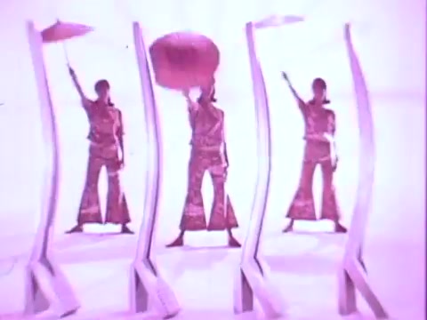 The still from the 'Adorn Hairspray Commercial' features three performers prominently dressed in vibrant outfits, striking dynamic poses. They hold large umbrellas above their heads against a bright pink backdrop. The styling and color palette convey a playful, retro aesthetic, typical of commercial visuals aimed at showcasing hair products. The overall composition emphasizes movement and flair in a lively setting.