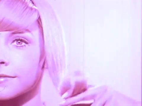 The still from the 'Adorn Hairspray Commercial' features a close-up of a young woman with straight, light-colored hair styled in a sleek manner. She has a subtle expression, and her hair is being lightly brushed or styled by a hand that is partially visible. The background has a soft, pinkish hue, contributing to a retro and vibrant aesthetic typical of vintage advertisements. The overall tone emphasizes beauty and hair care.
