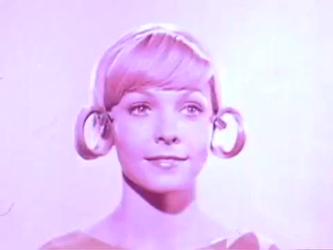 The still features a woman with a smooth, blonde hairstyle styled into large, circular rolls on either side of her head. She has a serene expression, with her gaze directed slightly upward. The background and color scheme are predominantly soft pink, creating a playful and vintage aesthetic typical of hair product advertisements from that era. This highlights the product, Adorn Hairspray, emphasizing the hairstyle's volume and hold.