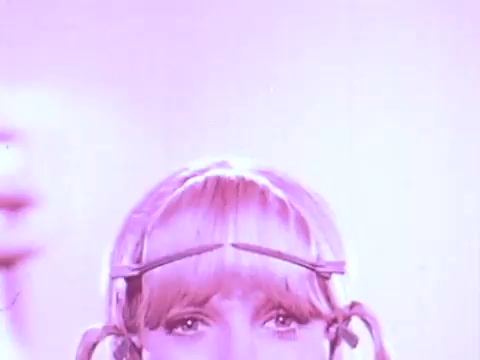 The still features a woman with light blonde hair styled in playful pigtails, wearing a headband that holds her hair back. She has a neutral expression and is framed against a soft pink background, which enhances the vibrant colors typical of 1960s aesthetics. The overall look suggests a retro, fun vibe, aligning with the context of a hairspray commercial aimed at showcasing hair styling solutions.