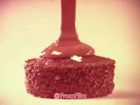 The still from the 'Hostess Ding Dong' commercial features a close-up shot of a chocolate-covered cake. Chocolate glaze is being poured over the top of the round, chocolate cake, highlighting its rich texture and indulgent appearance. The warm tones and soft focus create a nostalgic and appetizing atmosphere, typical of vintage food advertising.