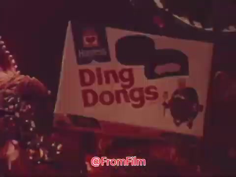 The still features a Hostess Ding Dongs box prominently displayed, likely amidst a colorful and festive backdrop typical of a commercial setting. The packaging is bold and eye-catching, highlighting the product name 'Ding Dongs.' The overall tone suggests a playful and inviting atmosphere.