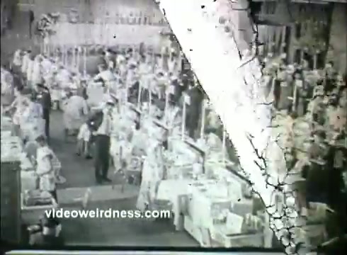 The still from 'Kaiser Aluminum - Pillsbury Bake Off 1957' showcases a bustling baking competition. An array of judges and competitors is visible, surrounded by tables laden with baked goods. The scene captures a sense of excitement and community, typical of a 1950s baking event. The image shows signs of wear typical of 16mm film, contributing to a nostalgic atmosphere.