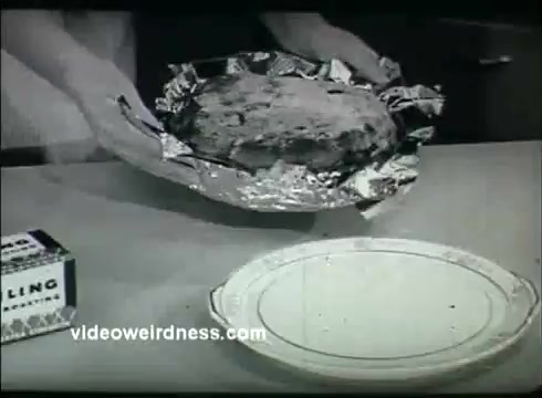 The still is from a 16mm film featuring a person's hands holding a baked item wrapped in aluminum foil. The scene includes a plate ready to receive the baked good and a box of Pillsbury baking mix positioned nearby. The overall tone is reminiscent of a mid-20th century cooking demonstration, emphasizing the use of Kaiser Aluminum materials in kitchen practices. The film captures a moment of anticipation in baking.