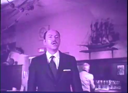 The still features Alan Arkin in a formal suit, displaying a surprised or contemplative expression. The setting appears to be a dimly lit bar or restaurant, with shelves holding glassware and a model ship adorning the wall in the background. The color tone of the image has a purple hue, typical of a 16mm print. The scene exudes a sense of tension or drama.