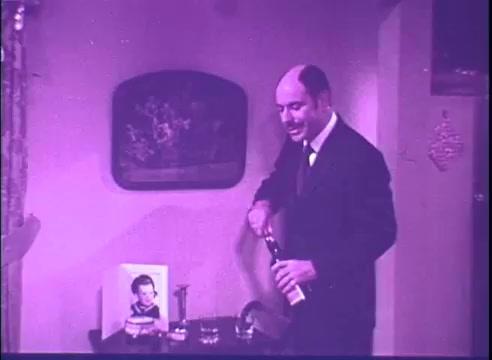 The still from the trailer of 'Last of the Red Hot Lovers' features a man in a suit, likely Alan Arkin, standing next to a table. He is in the process of opening a bottle, with a cigar and some drinks visible on the table. In the background, there is a painting on the wall and a framed picture of a young boy. The overall scene has a slightly washed-out purple tint, characteristic of a 16mm print.