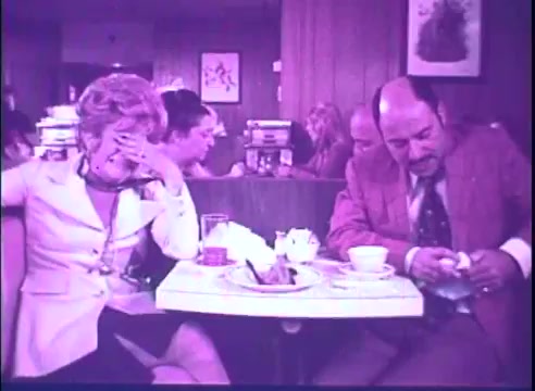 In this still from the trailer for 'Last of the Red Hot Lovers,' we see a tense scene set in a restaurant. A man, dressed in a suit, sits at a table, looking down and appearing troubled or contemplative. Opposite him, a woman in a light-colored outfit seems distressed, covering her face with her hand. The ambiance suggests a mix of complexity and discomfort, characteristic of the film's comedic yet poignant exploration of relationships. The color tone hints at the vintage 16mm film quality.