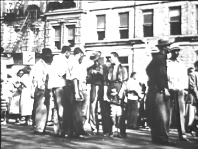 The still from the 'Ad Council PSA - Presidents Council on Youth Opportunity' depicts a group of individuals, primarily men, standing in a line on a city street. They appear to be in casual clothing, with some wearing hats. The setting features brick buildings in the background, indicating an urban environment. The scene conveys a sense of community or organization, possibly related to youth opportunities or initiatives. The overall tone seems historical, reflecting a moment from the past.