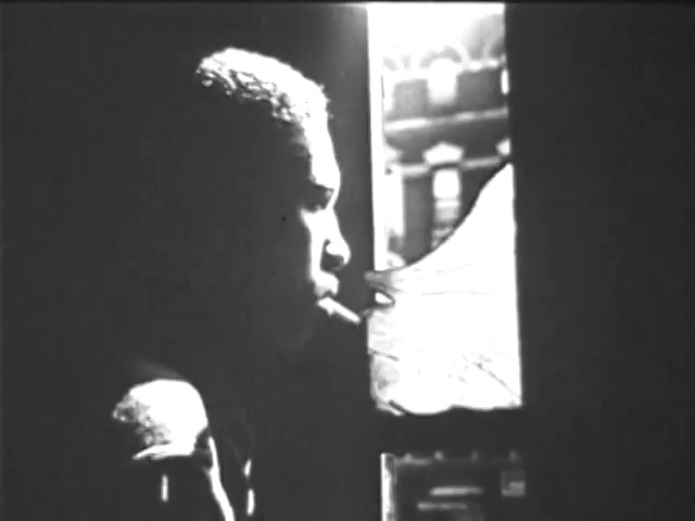 The still features a young man in profile, partially illuminated by light from a nearby window. He appears contemplative, holding a cigarette in his mouth. The contrast between light and shadow emphasizes his expression and the defining details around him. The scene suggests a moment of reflection or introspection, encapsulated in a vintage aesthetic typical of early film.