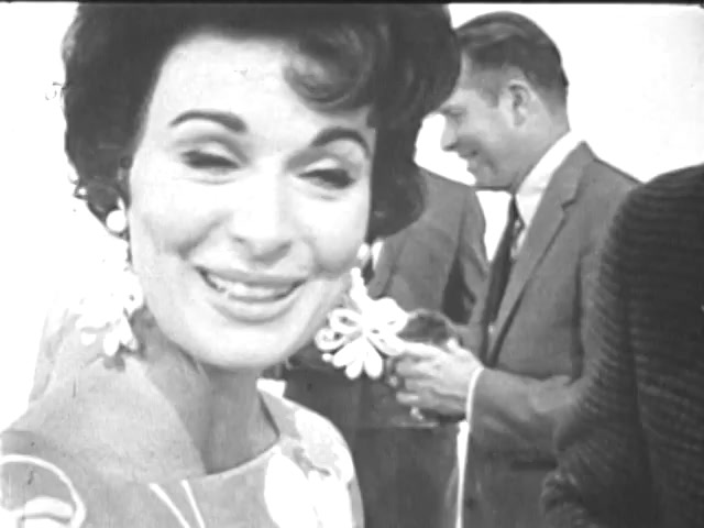 The still shows a woman with styled hair and large earrings smiling warmly, suggesting a friendly atmosphere. She appears to be engaged in conversation, with her expression conveying enthusiasm or joy. In the background, a couple of men in suits are visible, one of whom is holding something small, indicating a social or promotional setting related to youth opportunities. The overall tone hints at positivity and community engagement.
