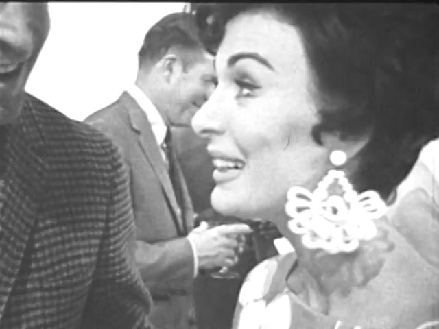 The still features a close-up of a woman engaging in conversation, smiling warmly. She wears distinctive large, decorative earrings and has a styled hairdo typical of the era. In the background, another man is slightly visible, dressed in a suit and tie, suggesting a social or formal setting. The overall tone is amiable and lively, indicative of a gathering focused on youth opportunities.